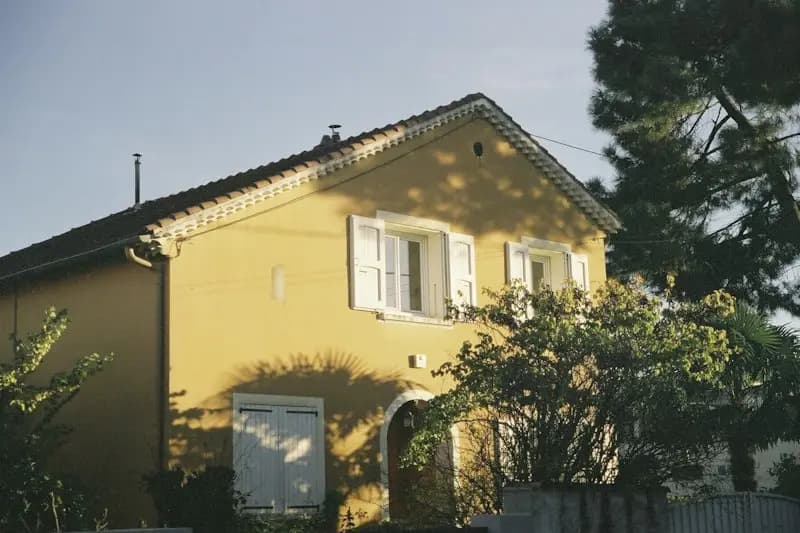 Maison à la façade jaune et aux volets blancs, dans un jardin arboré